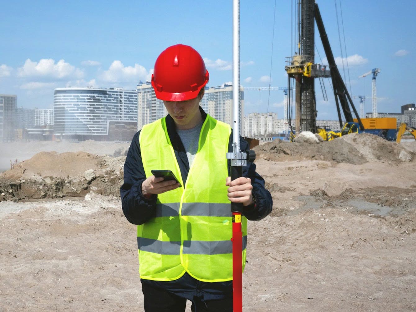 Site engineer in a red hard hat and hi-vis vest holding a survey pole on an open groundworks site with a piling rig behind